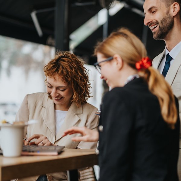 Three professionals engaging in a collaborative discussion while working on a laptop outdoors at a cafe. Represents teamwork, brainstorming, and productivity in a relaxed business environment.