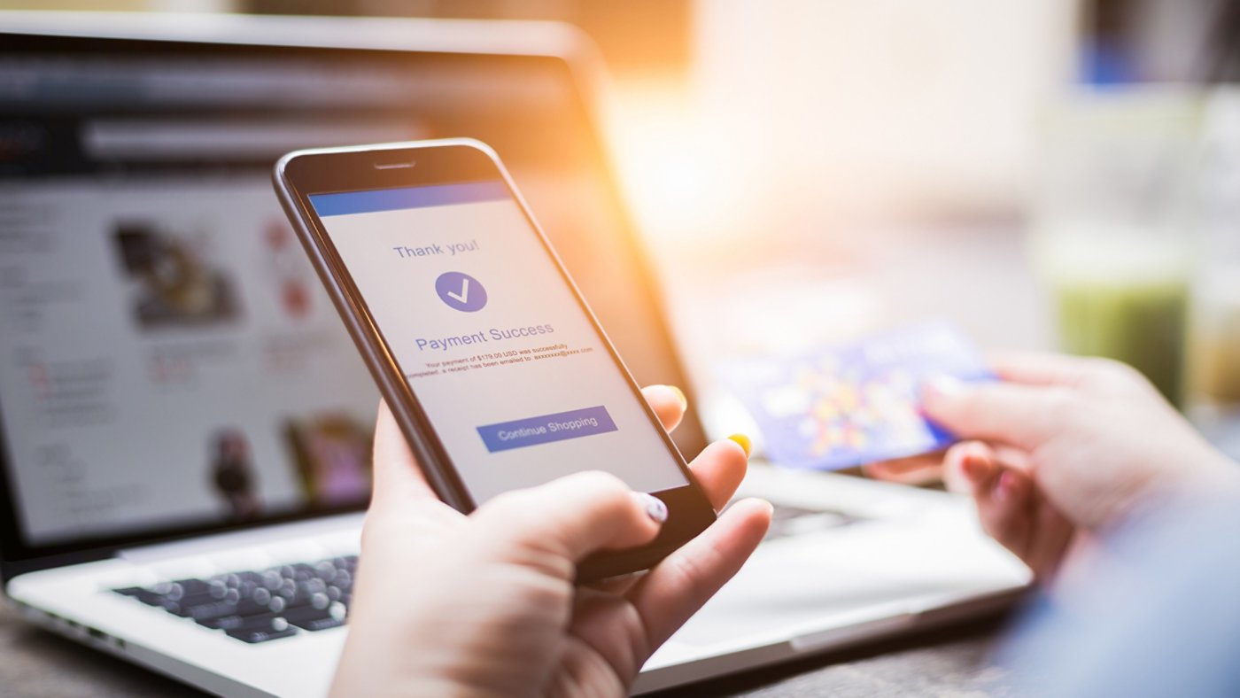 Online shopping concept, young woman hands holding mobile phone showing payment success information on screen with credit card and laptop computer on table while relax at home