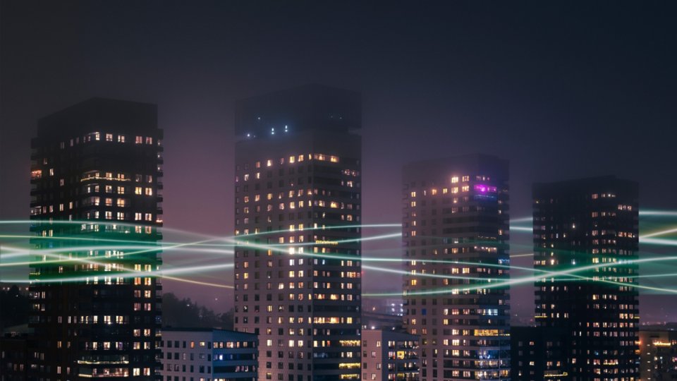 Tall residential or office towers rise against a dark night sky, their windows glowing with warm interior lights. A hazy, purple-toned atmosphere surrounds the cityscape, suggesting fog or light pollution. Bright, flowing light trails sweep horizontally across the scene, evoking digital connectivity or high-speed data. The overall mood is modern and futuristic, blending urban architecture with technology-inspired visuals.