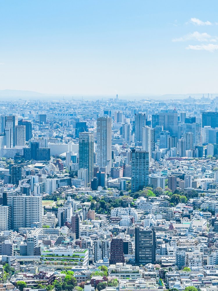 A wide-angle view of a bustling cityscape featuring modern skyscrapers and densely packed buildings. The setting is under a clear blue sky with scattered clouds, creating a bright and vibrant atmosphere. Green spaces are visible amidst the urban structures, adding contrast to the concrete environment.