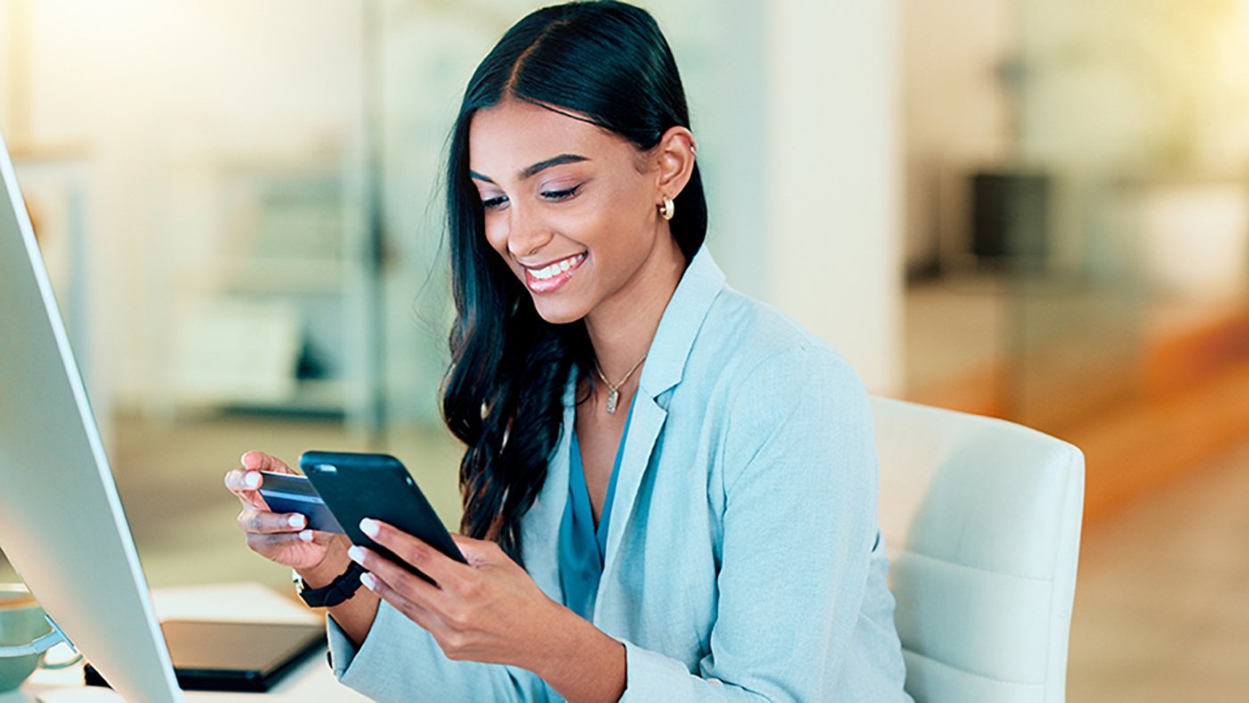 Happy woman making online payment with phone and bank card for typing credit details into online banking app. Young business woman enjoying virtual shopping or wireless purchase while sitting inside.