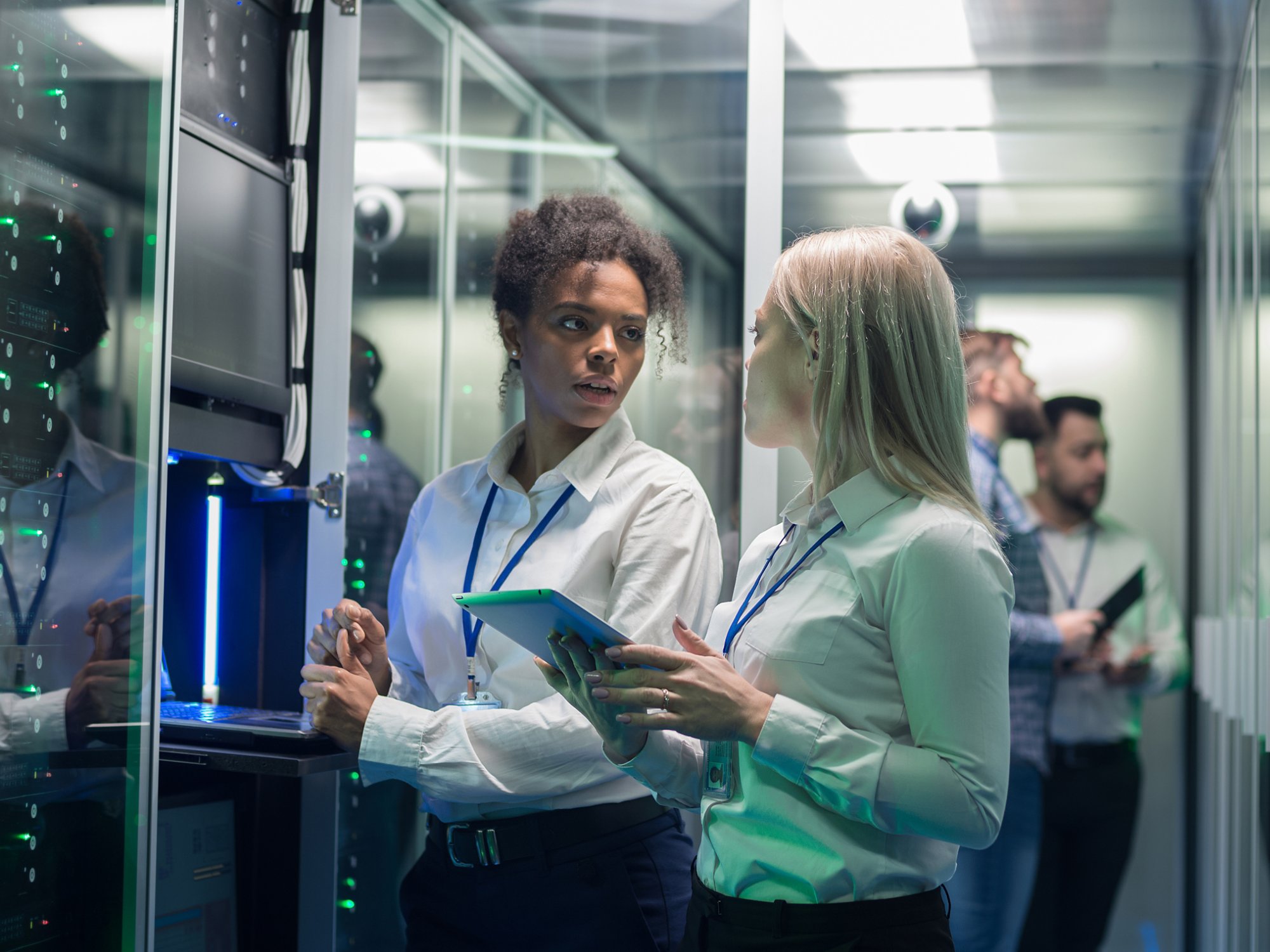 Medium shot of two women working in a data center with rows of server racks and checking the equipment and discussing their work