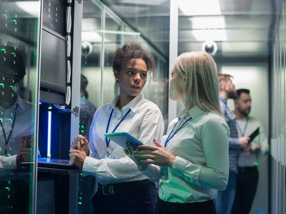 Medium shot of two women working in a data center with rows of server racks and checking the equipment and discussing their work