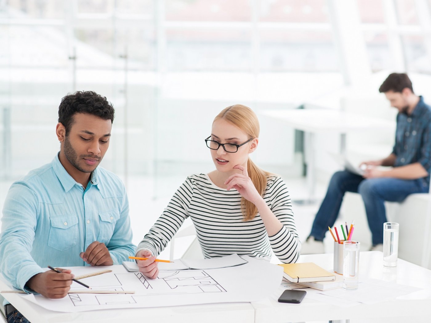 Be part of the solution. Two young architects sitting at white table and discussing drawing with their colleague working in background