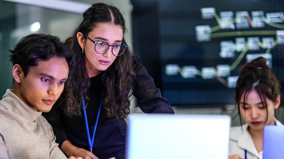 A diverse team of AI developers collaborates on a night coding session. A senior engineer oversees his team, intensely focused on debugging a complex machine learning project.