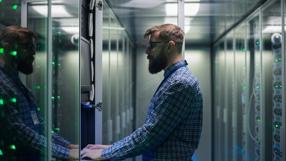 Adult bearded system administrator in glasses working on laptop and correcting errors in server hardware