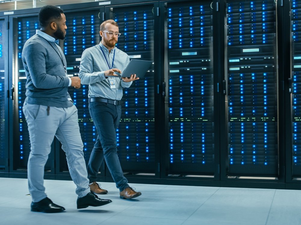 IT Technician with a Laptop Computer and Black Male Engineer Colleague are Talking in Data Center while Walking Next to Server Racks. Running Diagnostics or Doing Maintenance Work.