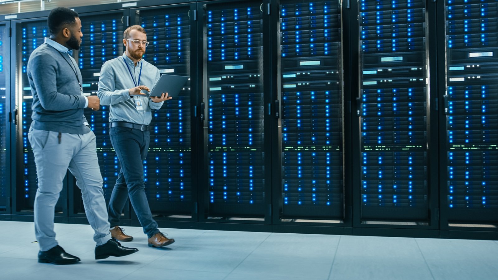 IT Technician with a Laptop Computer and Black Male Engineer Colleague are Talking in Data Center while Walking Next to Server Racks. Running Diagnostics or Doing Maintenance Work.