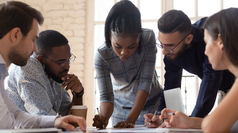 Focused african american millennial female team leader leaned over table, writing down project ideas, editing documentation at brainstorming business meeting with diverse partners or clients at office.