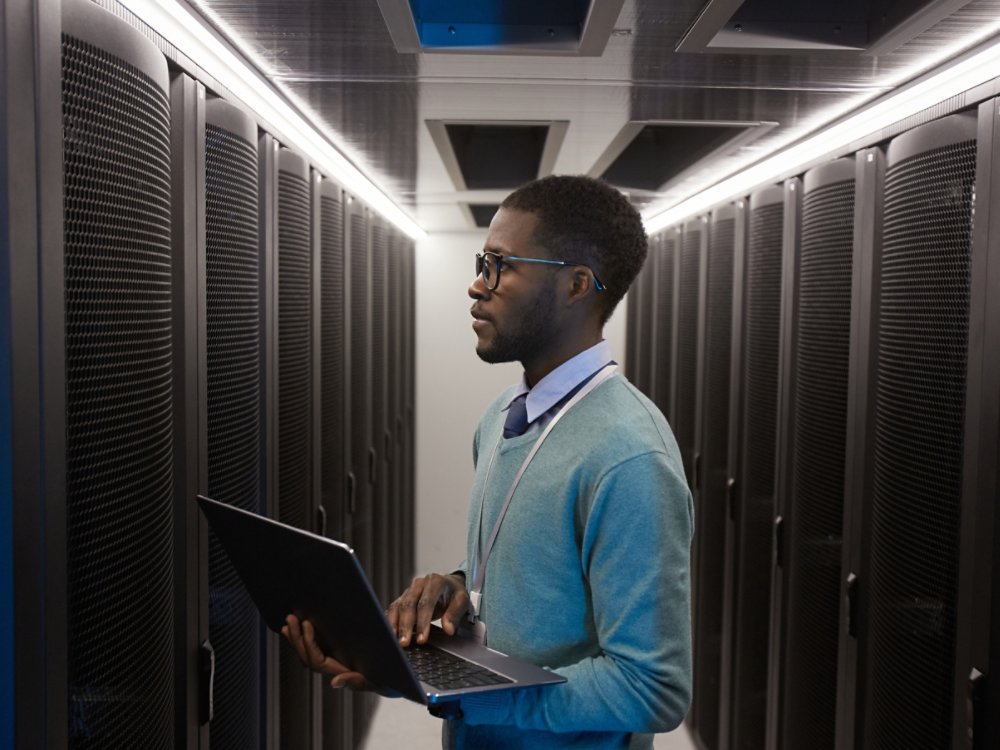 Side view portrait of young African American data engineer working with supercomputer in server room and holding laptop, copy space