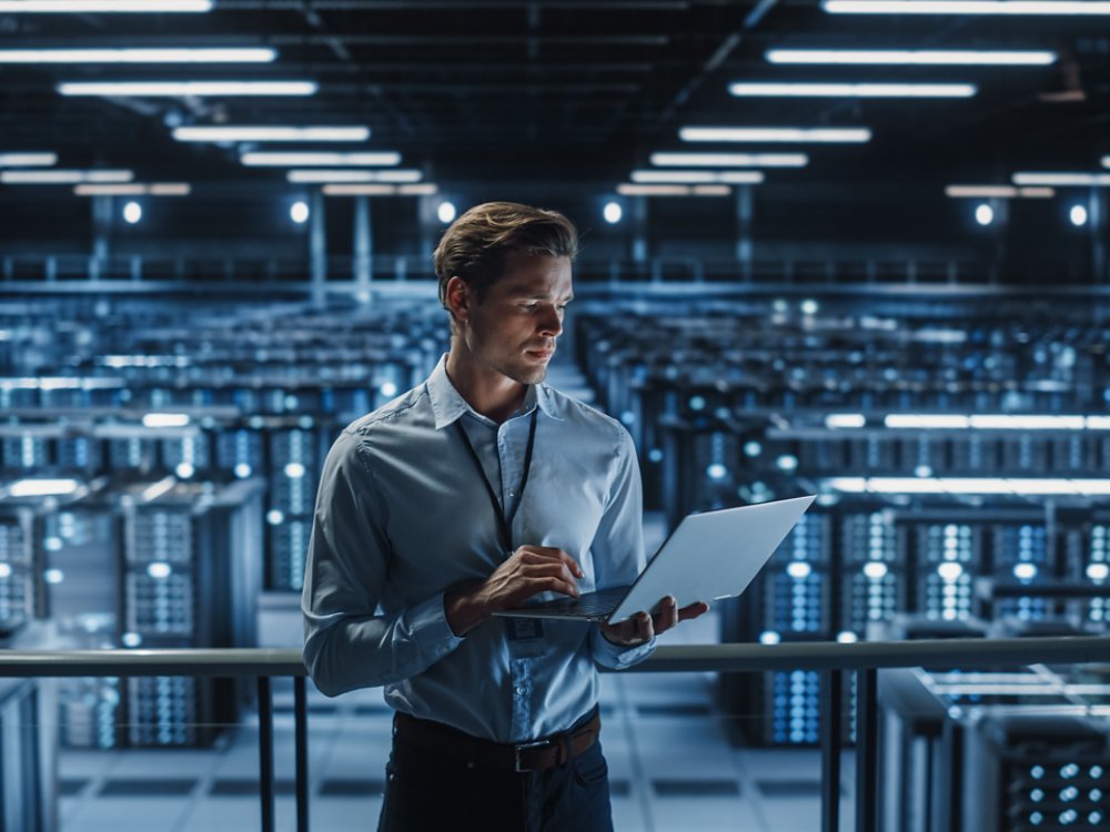 Portrait of IT Specialist Uses Laptop in Data Center. Server Farm Cloud Computing Facility with Male Maintenance Administrator Working on Cyber Security and Network Protection.