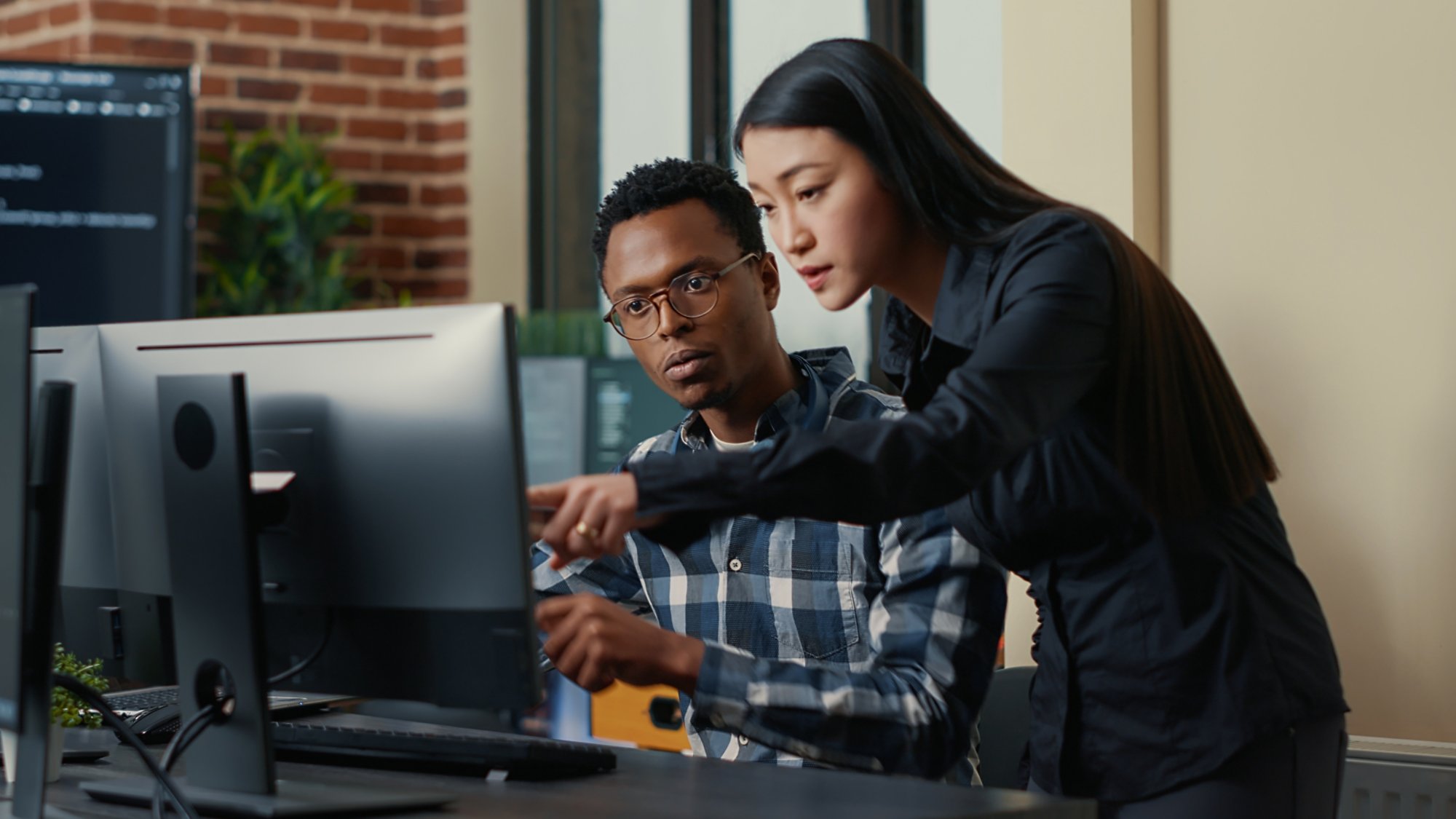 aTwo software developers coming at desk and sitting down holding laptop with coding interface and pointing at computer screen. Programmers team discussing algorithms looking at monitors with code.
