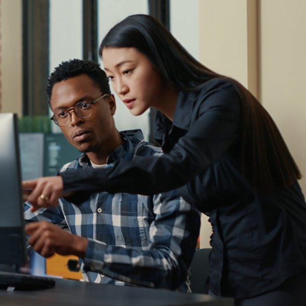 aTwo software developers coming at desk and sitting down holding laptop with coding interface and pointing at computer screen. Programmers team discussing algorithms looking at monitors with code.