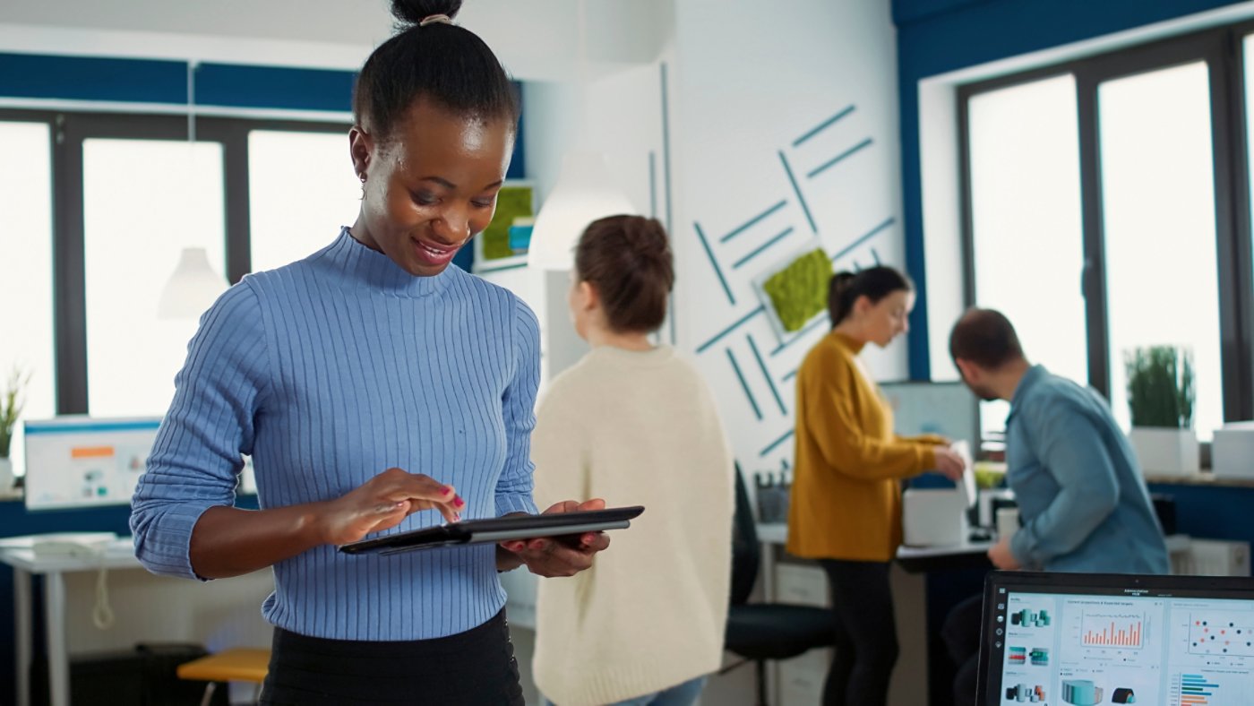 Portrait of african american woman standing in busy office picking up tablet with business erp software and smiling