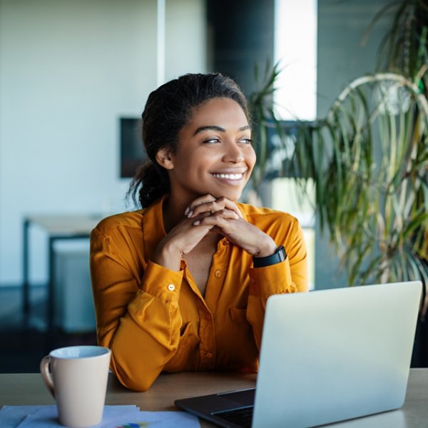 Portrait of dreamy african american businesswoman sitting at desk in office and thinking while working on laptop, looking away with pleased face expression, copy space