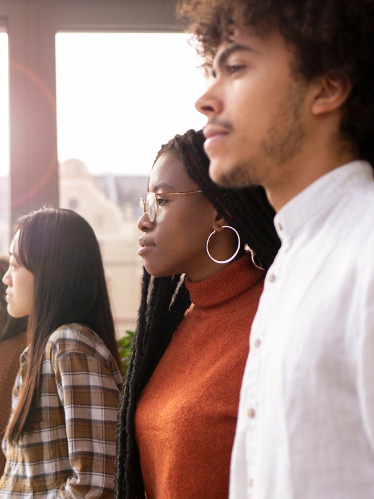 A diverse group of individuals stands indoors, gazing through a large glass window. The setting appears to be modern and well-lit, with reflections visible on the glass. The group is dressed in casual attire, featuring warm tones and plaid patterns.