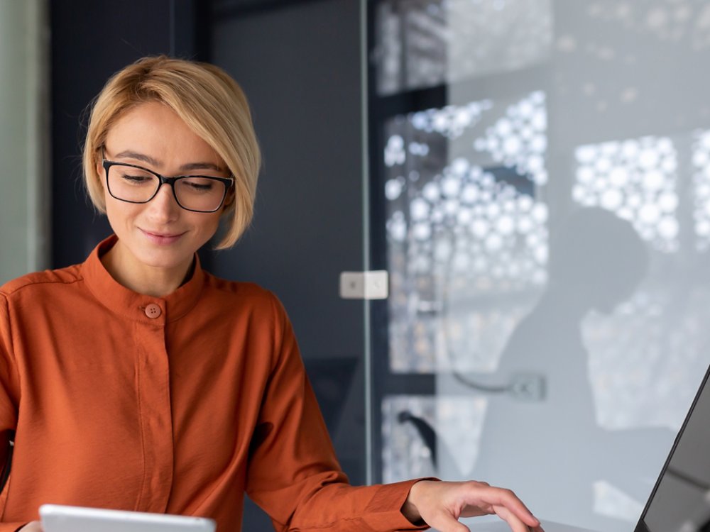 Young successful businesswoman at workplace is satisfied with results of achievement at work smiling and using tablet computer, blonde programmer is testing new software in application.