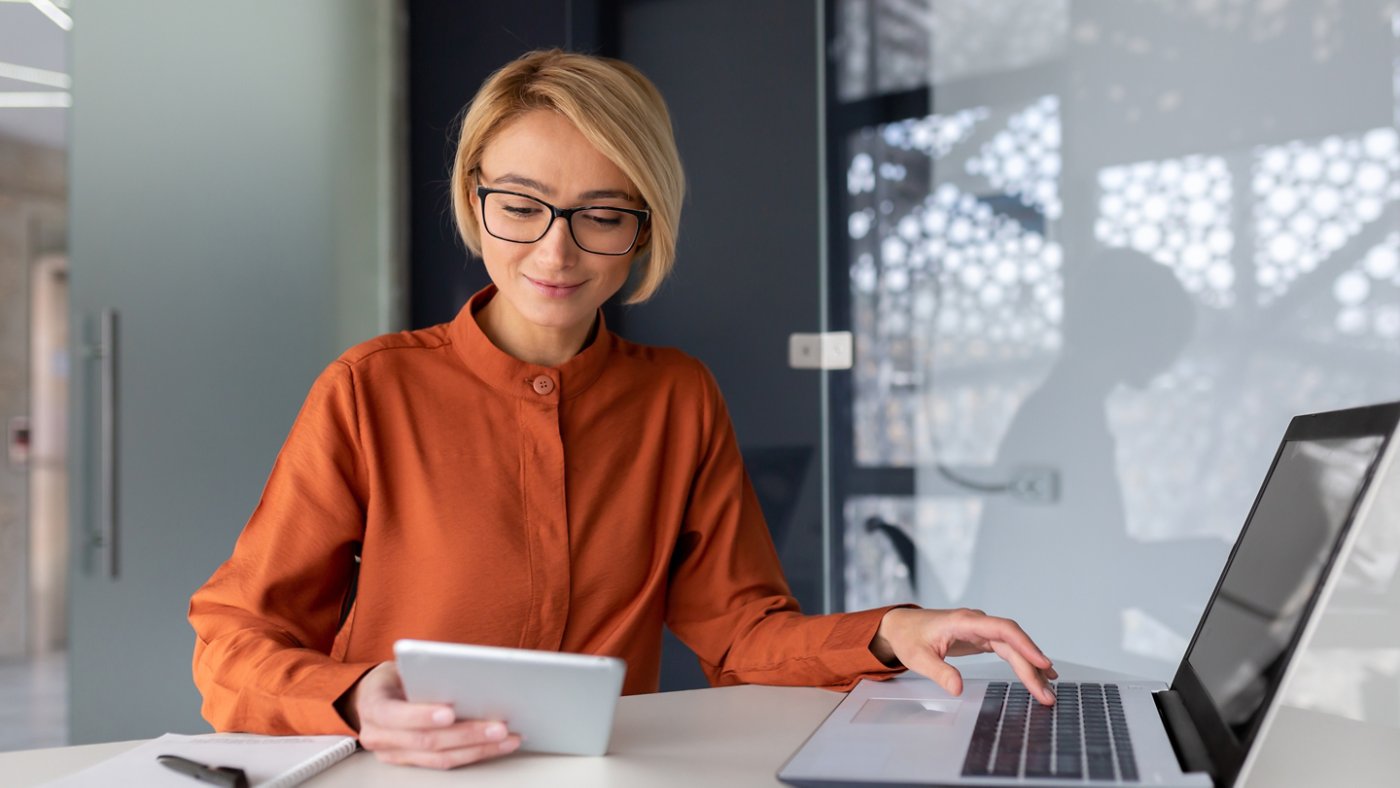 Young successful businesswoman at workplace is satisfied with results of achievement at work smiling and using tablet computer, blonde programmer is testing new software in application.