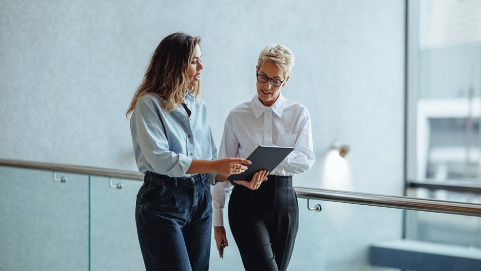 Mature business woman looking at a tablet screen as her assistant discusses her schedule with her. Two professional business women planning meetings and appointments in an office.