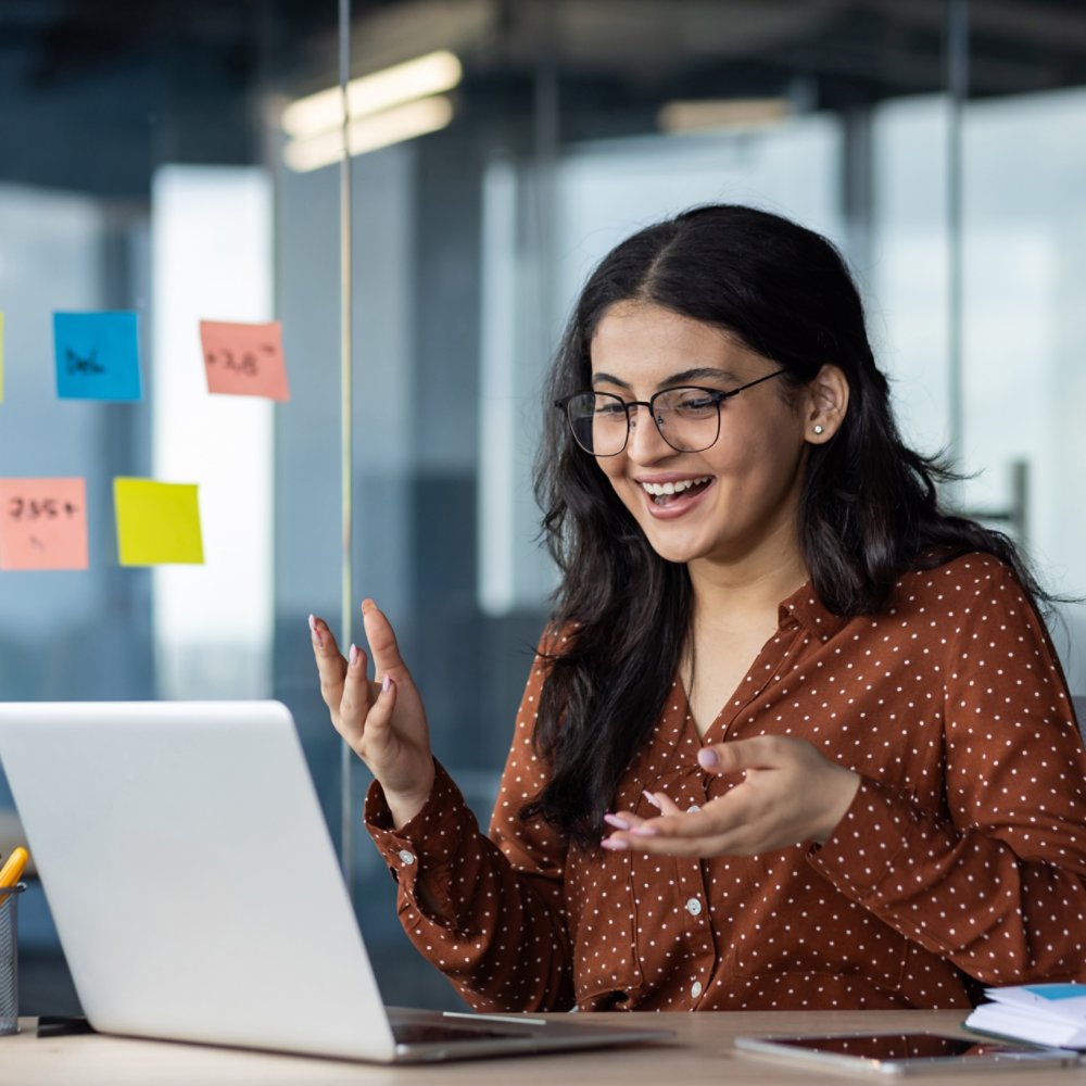 Young business woman inside office using laptop for video call, female worker smiling and talking with colleagues remotely in online meeting, joyful female worker with computer.