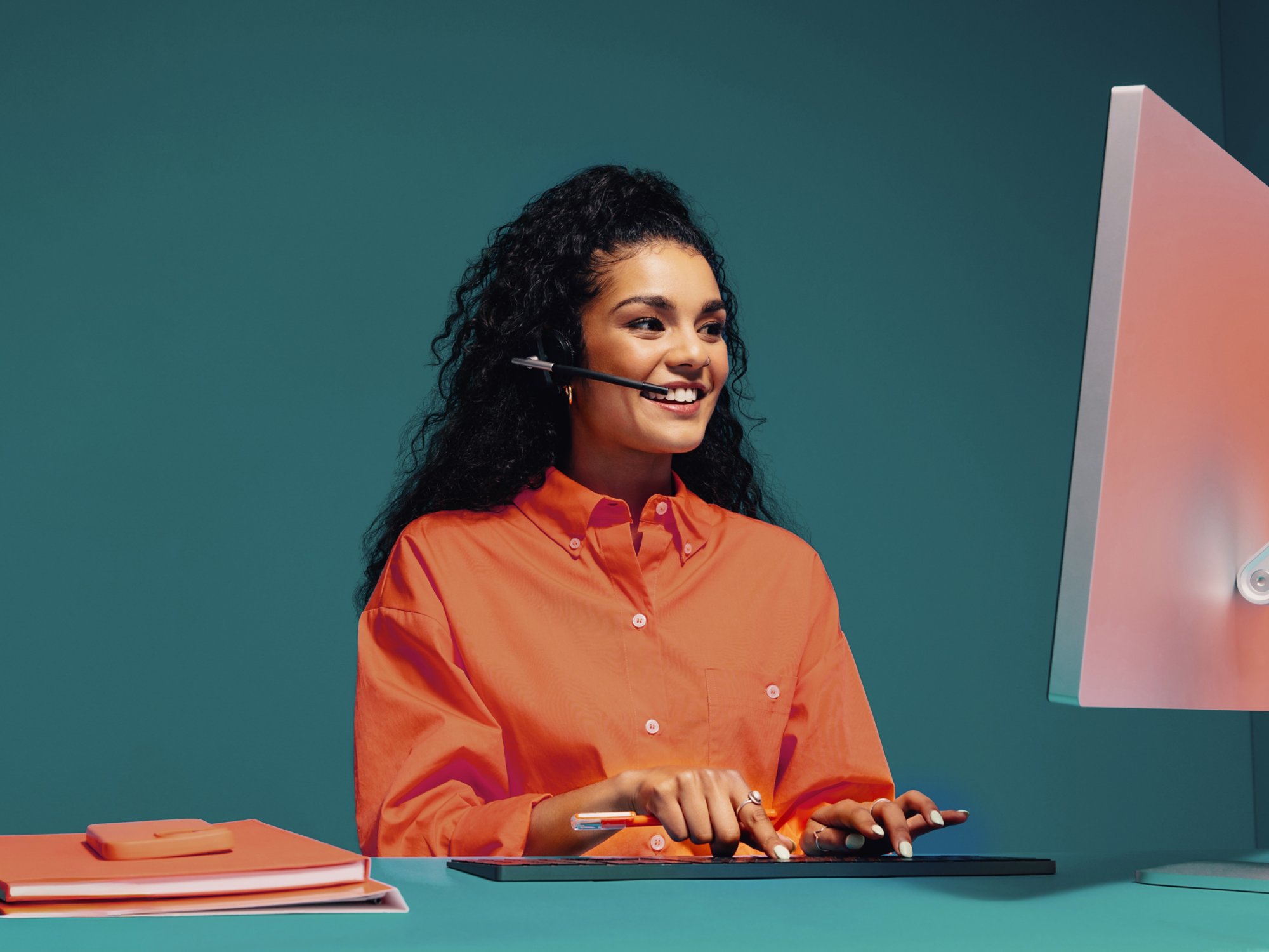 Smiling female agent wearing headset and typing on keyboard while working on a computer in a monochromatic office with bright orange elements.