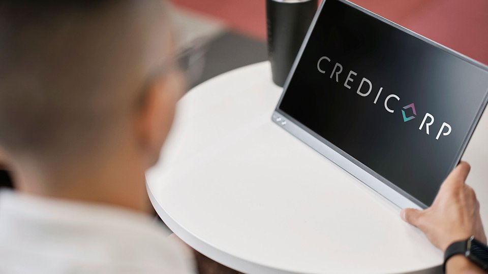 A person sits at a round white table viewing a tablet screen that displays the word CREDICORP. The scene appears to be in a modern office or workspace with a blurred background. A dark beverage cup is visible on the table, adding to the casual professional setting. The focus is on the digital device and the Credicorp text on the display.
