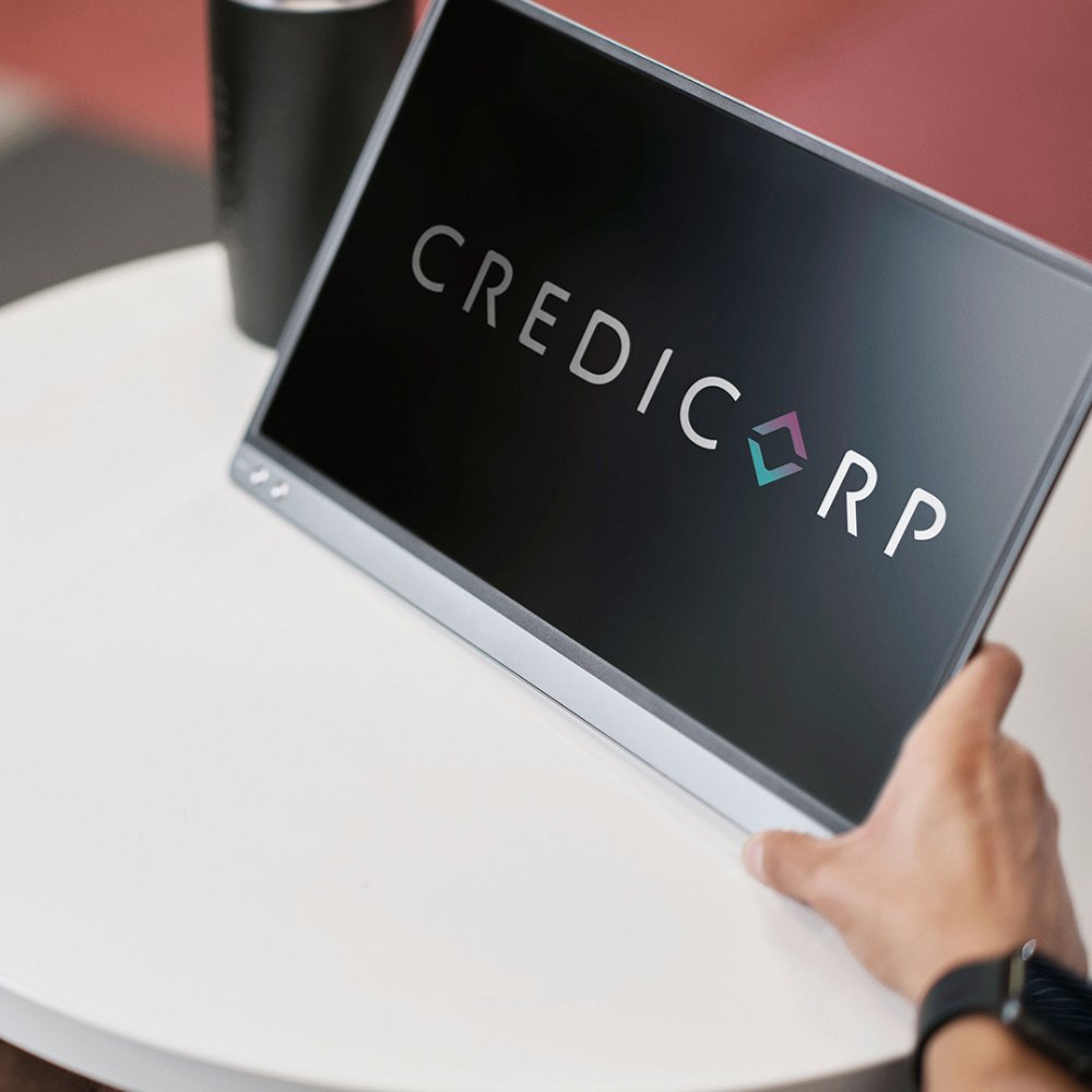 A person sits at a round white table viewing a tablet screen that displays the word CREDICORP. The scene appears to be in a modern office or workspace with a blurred background. A dark beverage cup is visible on the table, adding to the casual professional setting. The focus is on the digital device and the Credicorp text on the display.