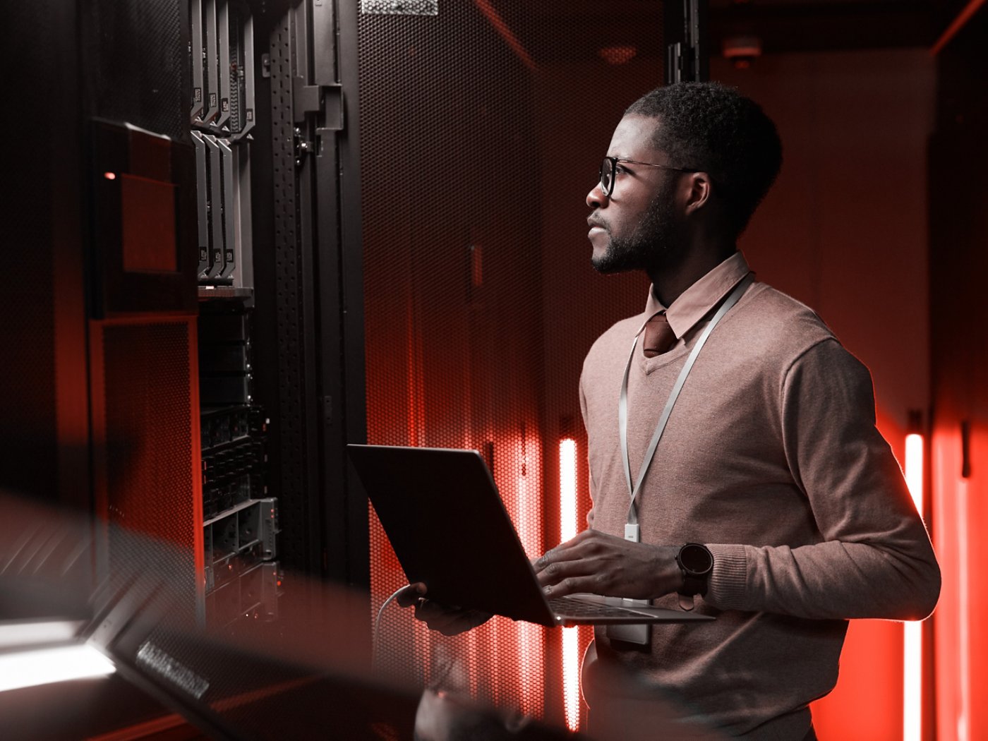 Side view portrait of African American data engineer holding laptop while working with supercomputer in server room lit by blue light, copy space