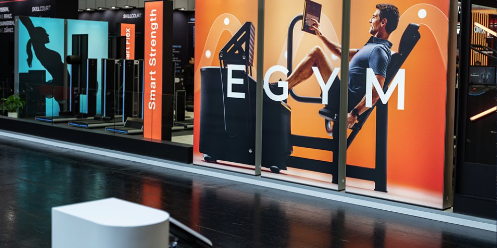 Large EGYM advertising panels showcase a man using a strength training machine while reading a tablet in a modern exhibition hall. The booth features bright orange and teal gradients with fitness silhouettes and equipment in the background. A vertical panel with the text "Smart Strength" highlights the focus on intelligent workout technology. The polished floor and clean structures emphasize a contemporary, professional trade show environment.