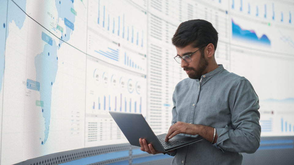 Indian Male Product Distribution Expert Holding Laptop Computer, Analyzing World Map On Big Digital Screen In Monitoring Office. Successful Man Looking For Efficient Routes For International Delivery.