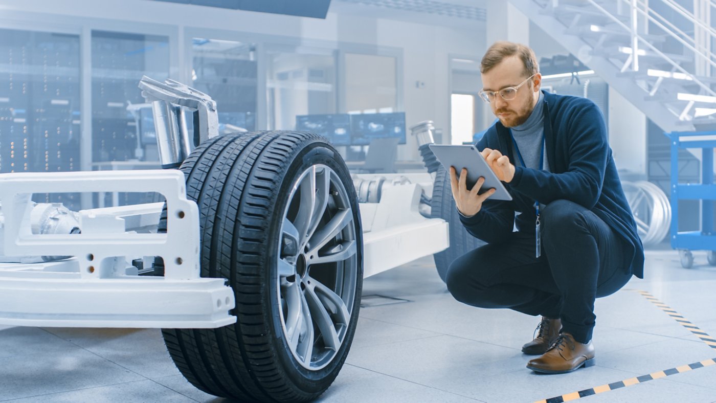 Engineer with Glasses and Beard Works on a Tablet Computer Next to an Electric Car Chassis Prototype with Wheels, Batteries and Engine in a High Tech Development Laboratory.