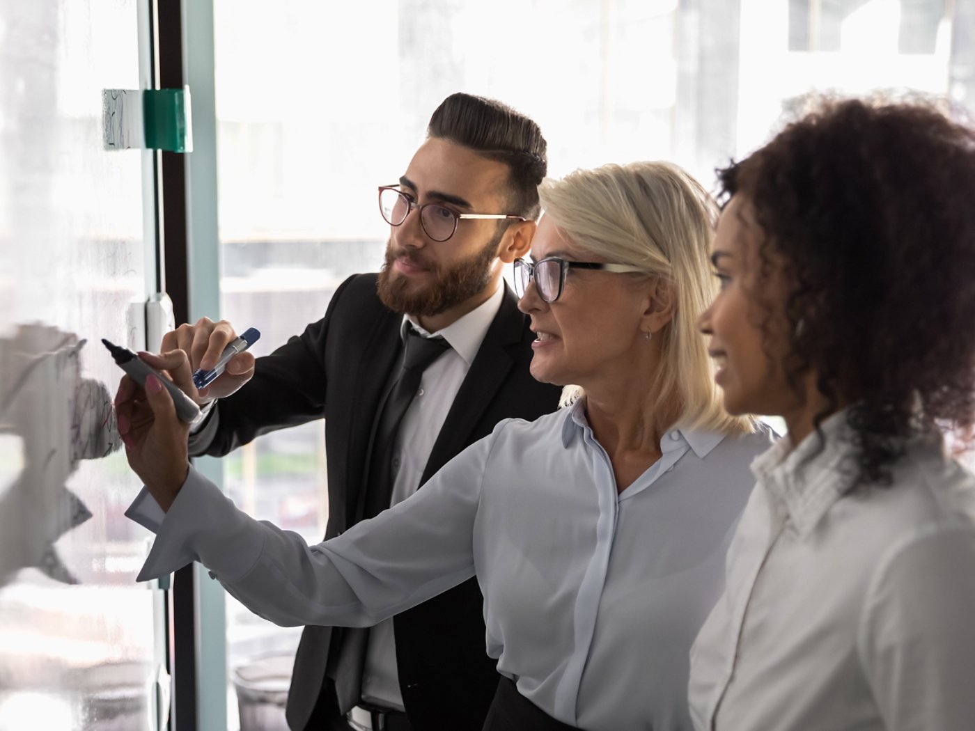 Serious focused multiracial businesspeople write draw on white board in office develop business project together, multiethnic colleagues engaged in creative thinking, brainstorm discussing strategy