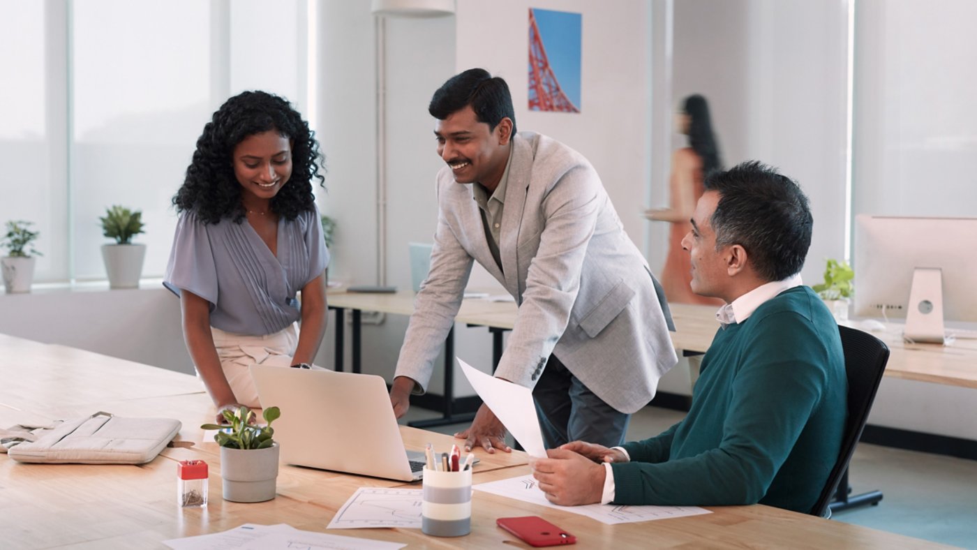 Three asian business people talking, with handheld report and a laptop on work-desk in office, with a lady walking by in the background 16X9