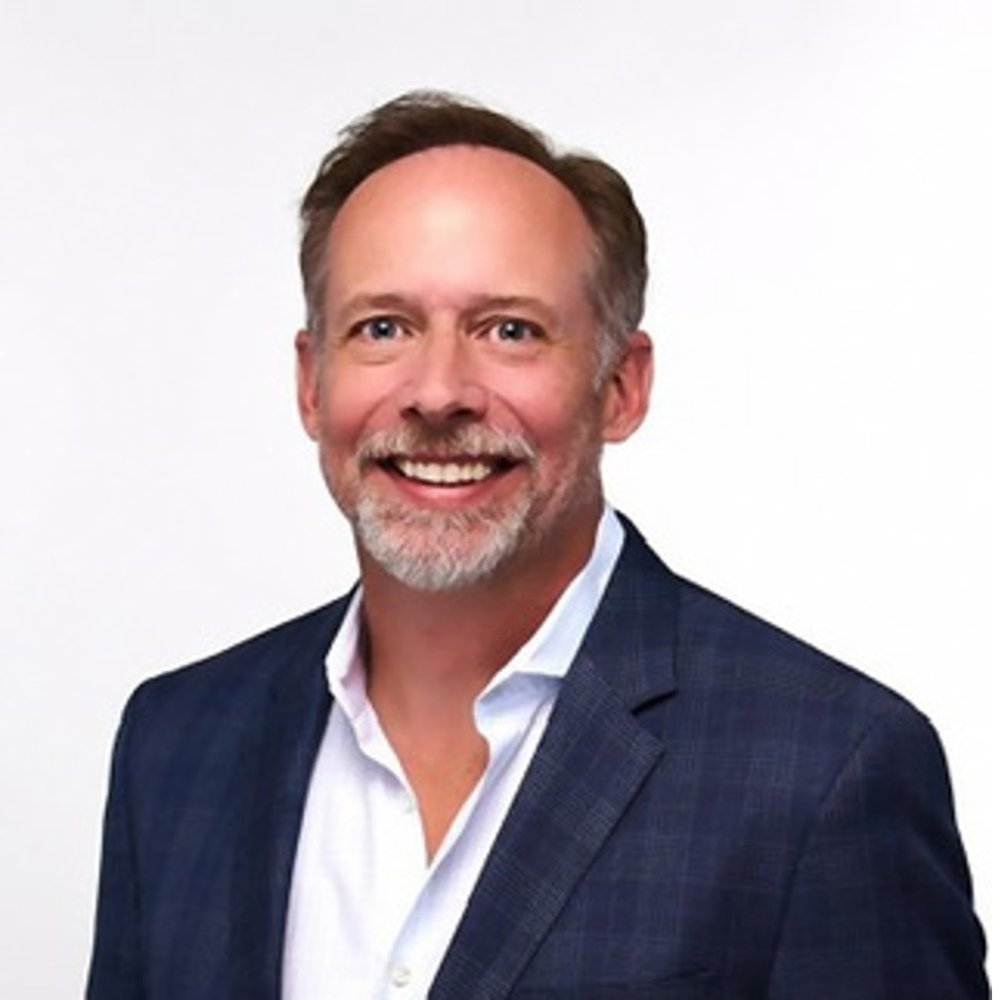 A man is dressed in a formal blue checkered blazer paired with a white collared shirt. The setting is a plain white background, emphasizing the professional attire. The blazer features a subtle checkered pattern, adding texture to the overall look.