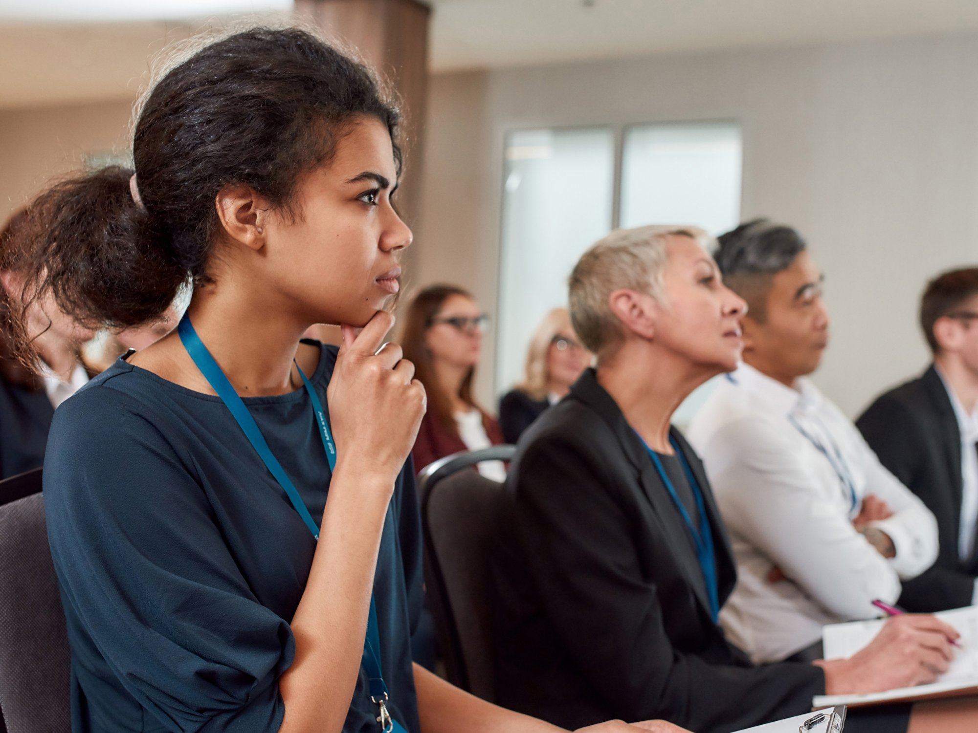 Multi-ethnic audience sitting in a row and making notes while listening to presentation at conference hall. Selective focus on woman. Horizontal shot. Side view