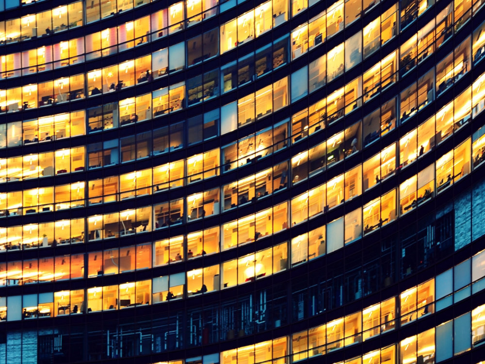 The image depicts workers working late in an office at night.  The employees are seen as silhouettes against their lit offices that have large windows.  The building is lit by the "blue hour" evening sky.  The building and its windows cover the entire canvas of the image.  It is apparent that the building may have some sort of curvature to the architecture.  While most of the building reflects patterns of yellow light, a small portion of it reflects the blue light.  There is no border around the image.  The windows on the edges and corners of the image are truncated in the view.