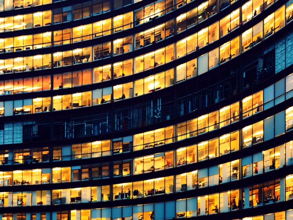 The image depicts workers working late in an office at night.  The employees are seen as silhouettes against their lit offices that have large windows.  The building is lit by the "blue hour" evening sky.  The building and its windows cover the entire canvas of the image.  It is apparent that the building may have some sort of curvature to the architecture.  While most of the building reflects patterns of yellow light, a small portion of it reflects the blue light.  There is no border around the image.  The windows on the edges and corners of the image are truncated in the view.