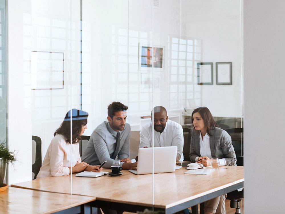 Diverse group of businesspeople working on a laptop while having a meeting around a table inside of a glass office boardroom