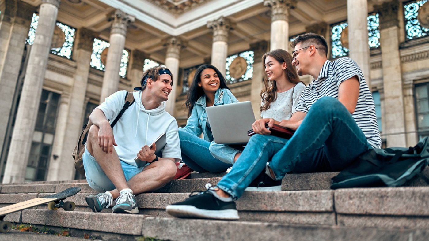 A group of students with laptops sit on the steps near the campus and communicate.