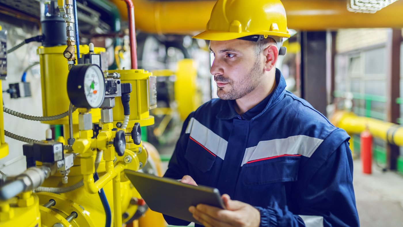 Serious caucasian unshaven worker in protective uniform and with hardhat using tablet for checking temperature in pipes. Factory interior.