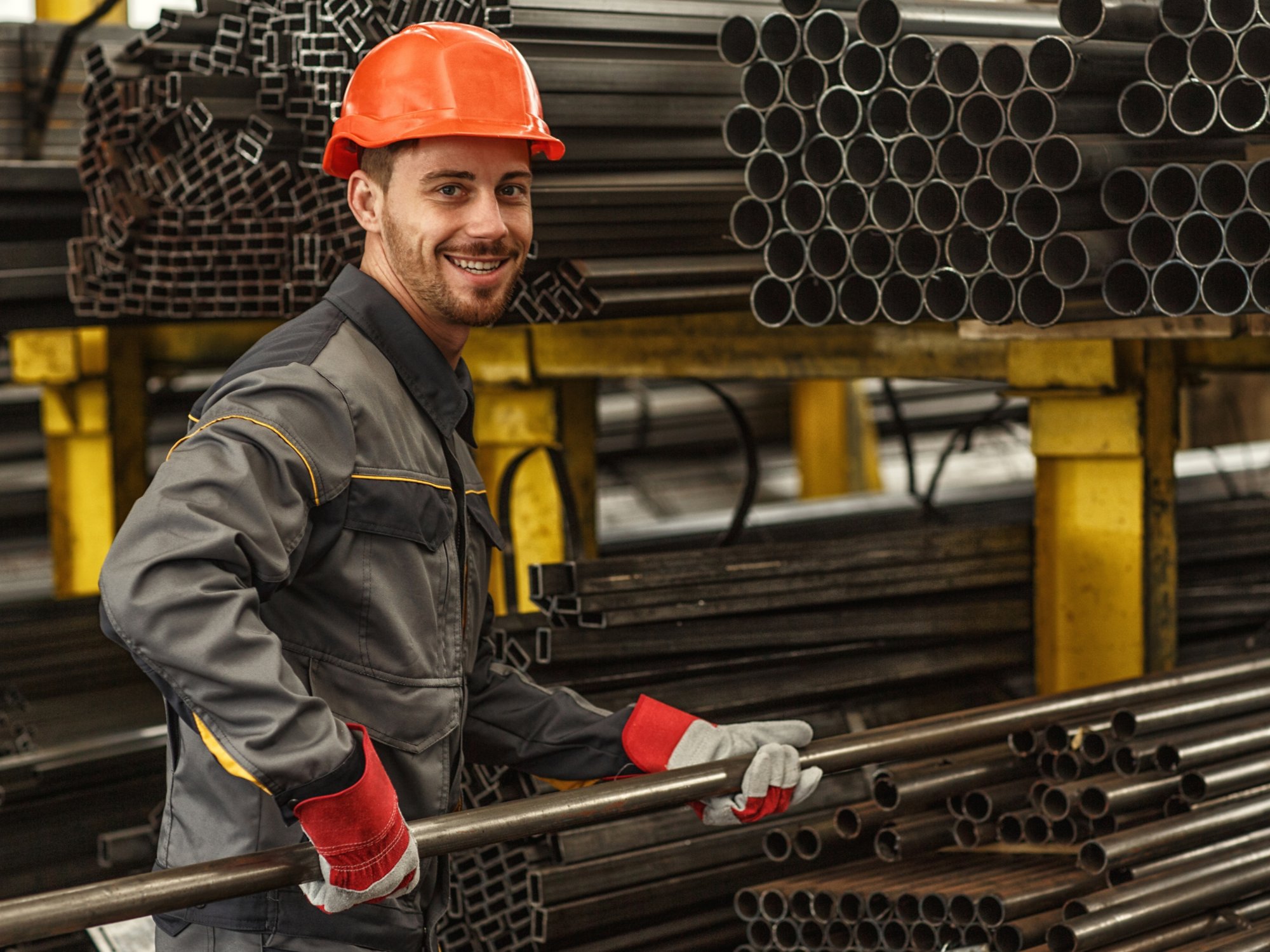 Horizontal portrait of a cheerful industrial worker in protective uniform and hardhat smiling to the camera over his shoulder while stacking steel pipes at the storage of a metalworking company