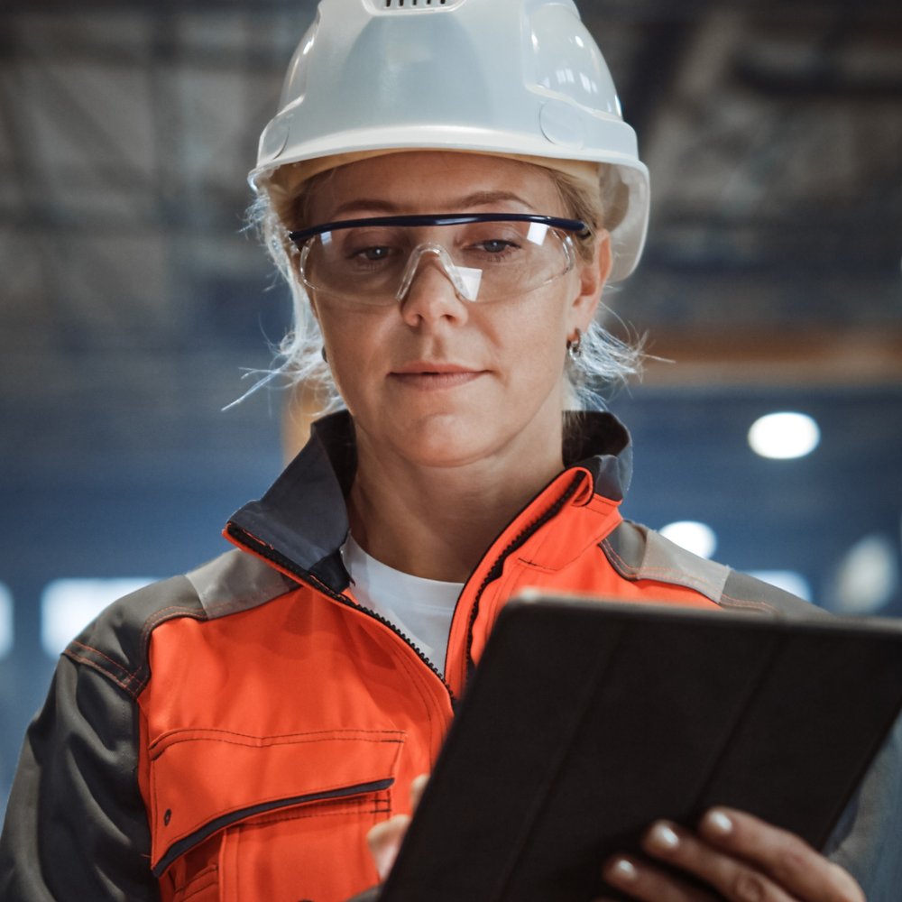 Professional Heavy Industry Engineer/Worker Wearing Safety Uniform and Hard Hat Uses Tablet Computer. Serious Successful Female Industrial Specialist Walking in a Metal Manufacture Warehouse.