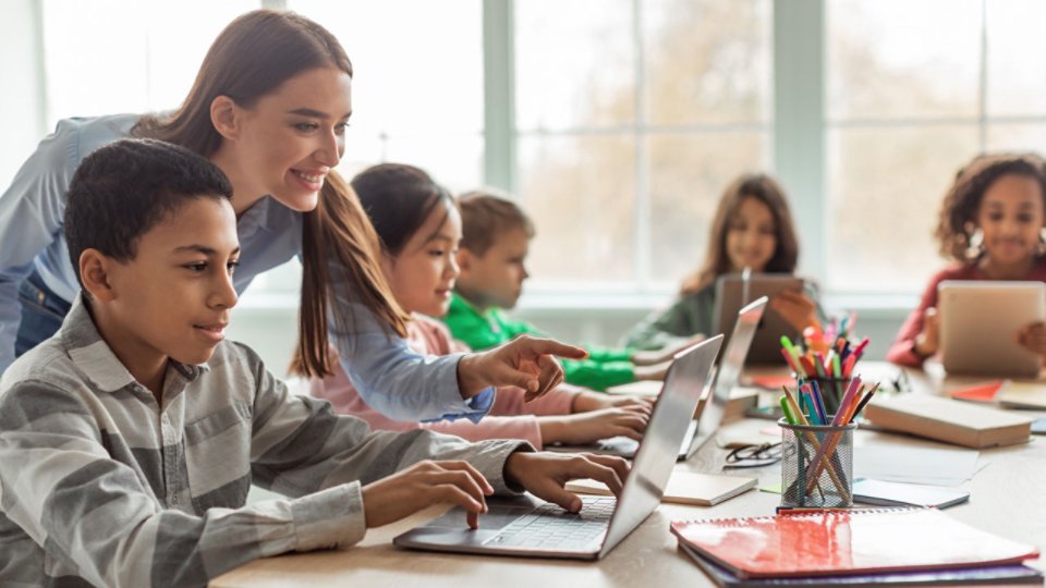 A teacher leans over to assist a student working on a laptop at a classroom table. Several children sit nearby using laptops and tablets, focused on their screens. The bright room features large windows and school supplies on the desk, suggesting a modern learning environment. The scene conveys collaborative education and the use of digital technology in school.