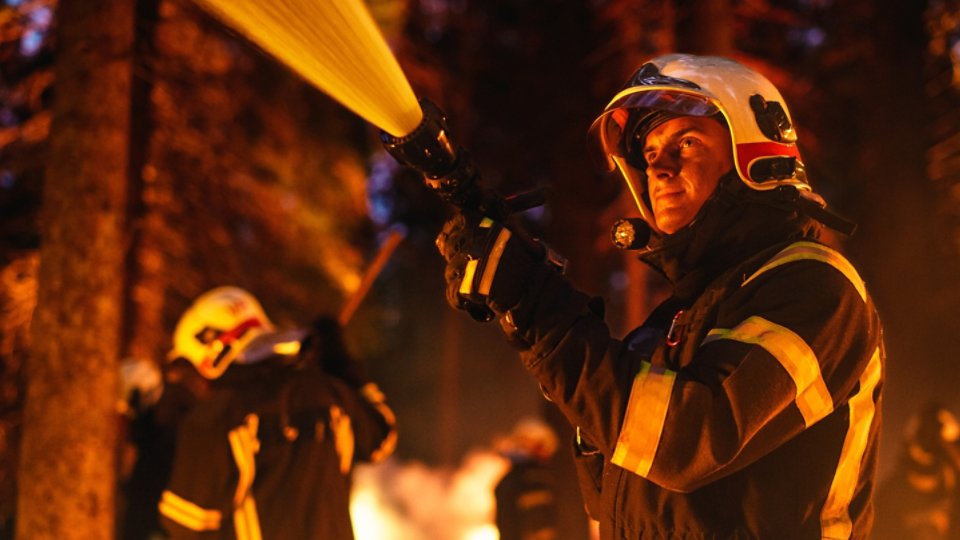 Portrait of a Handsome Caucasian Professional Firefighter Methodically Extinguishing a Forest Fire with a Fire Hose. Firemen Brigade Rescuing Wildland from Uncontrollable Arson.