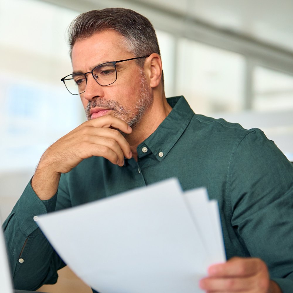 Busy middle aged executive, mature male hr manager holding documents using laptop looking at pc in office at desk