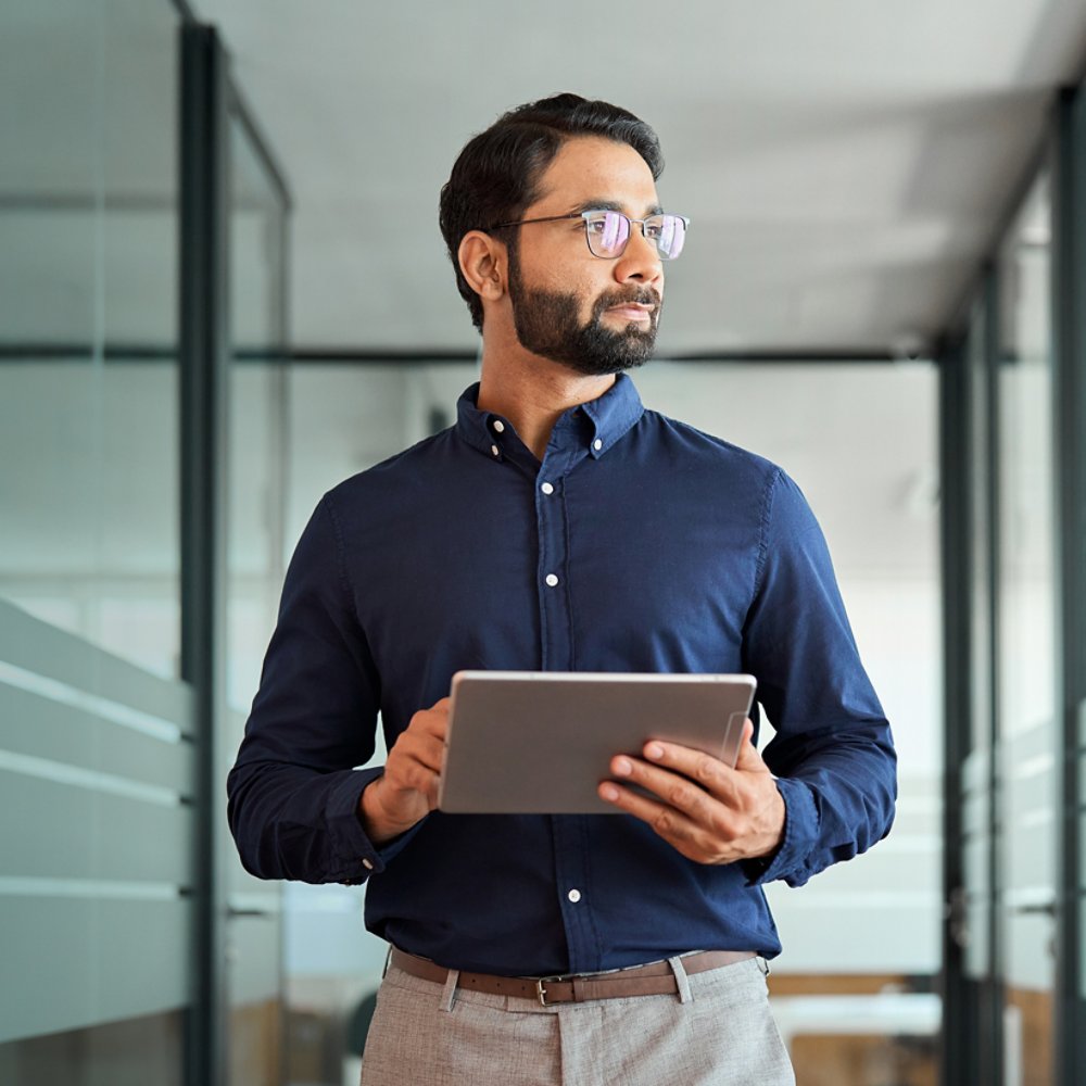 MAn walking down an office hallway with glass walls holding an computer tablet