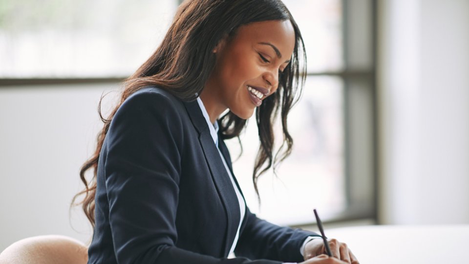 Smiling young African American businesswoman writing down notes in her day planner while sitting at a table in an office