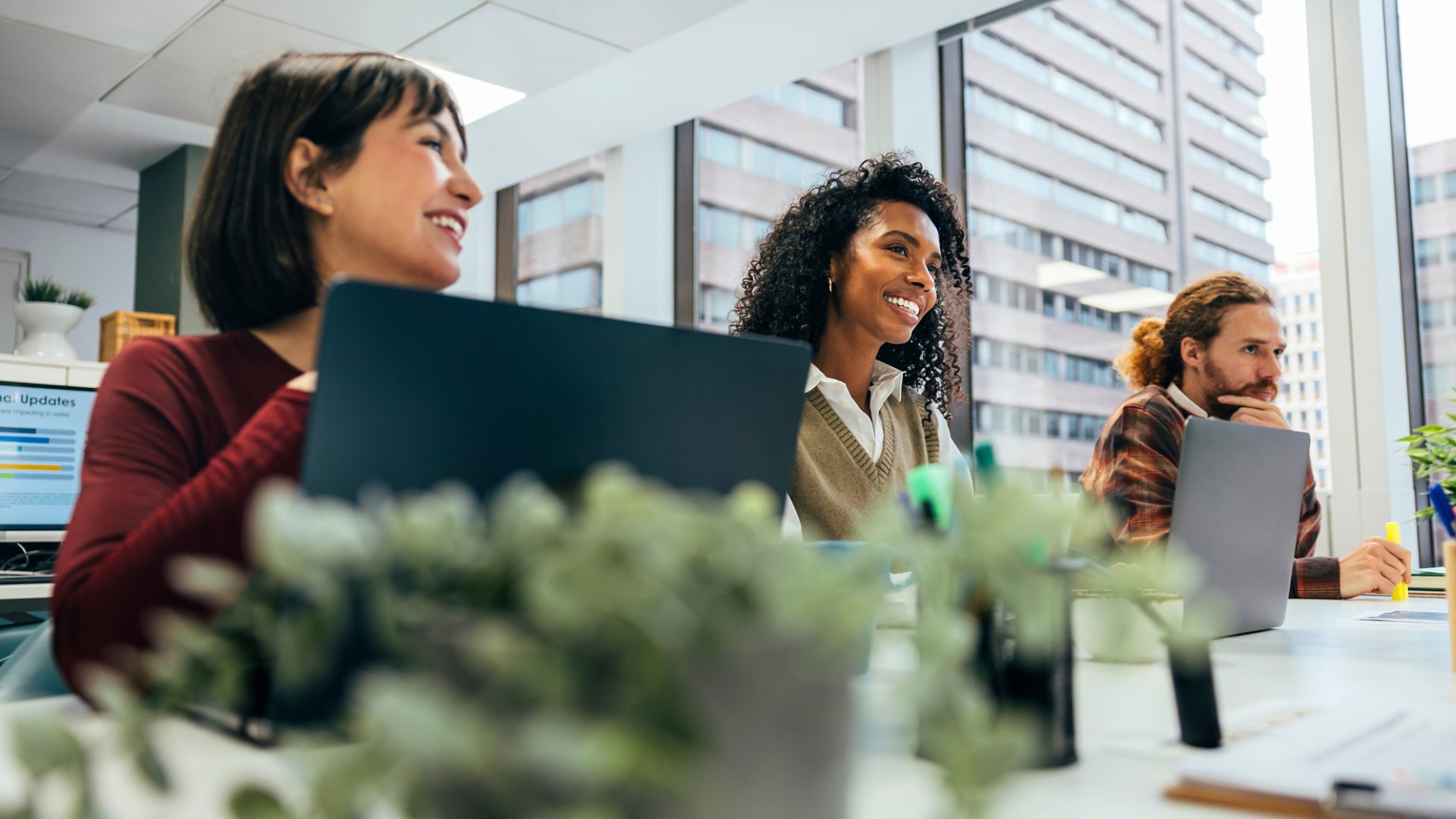 A young, diverse group of professionals engages in work at a well-lit office space with laptops, smiling and interacting in a collaborative environment.
