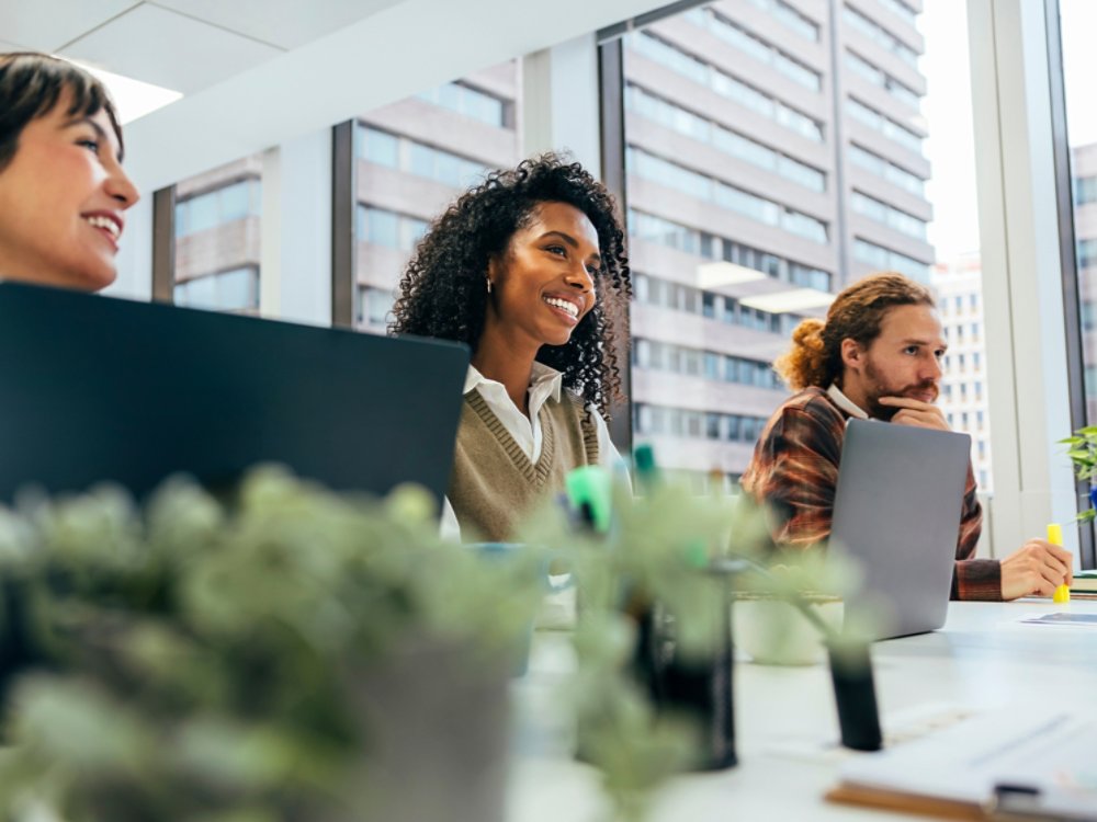 A young, diverse group of professionals engages in work at a well-lit office space with laptops, smiling and interacting in a collaborative environment.