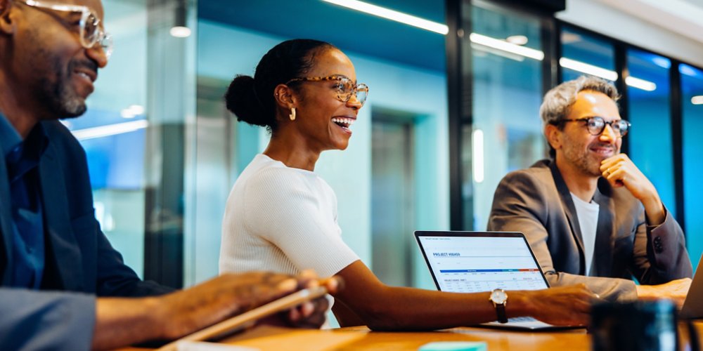 Three diverse coworkers, smiling and engaged in discussion around a meeting table in an office setting, collaborating with laptops and notes.